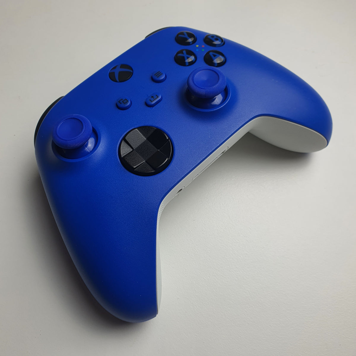 Front of a 'Shock Blue' Xbox game controller showing the front panel, including dual analog sticks, black directional and action buttons, and the Xbox logo, on a light white background.