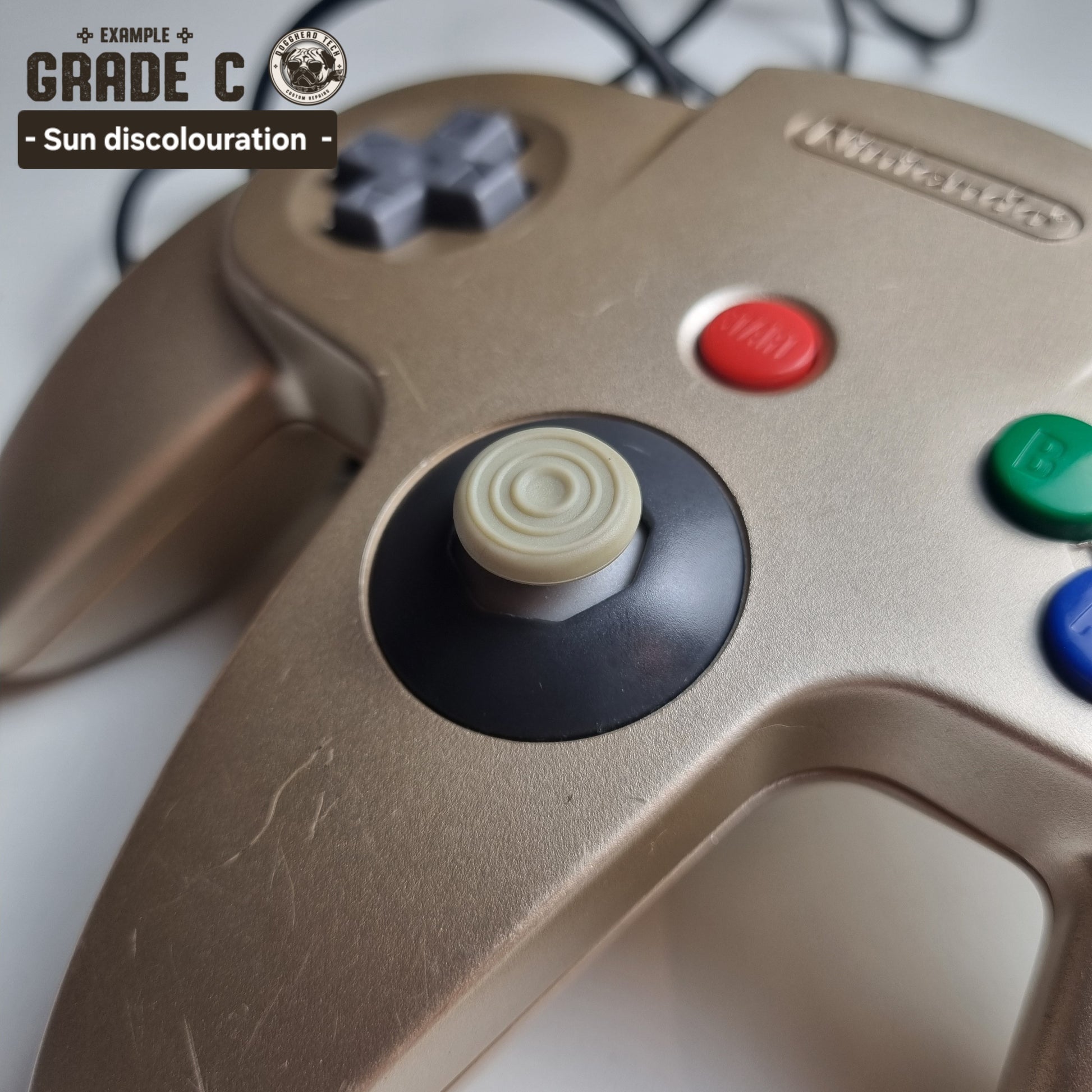 Close-up of the analog stick of a refurbished modified gold Nintendo 64 controller, showing some sun damage. The text reads 'Example Grade C: Sun discolouration'.