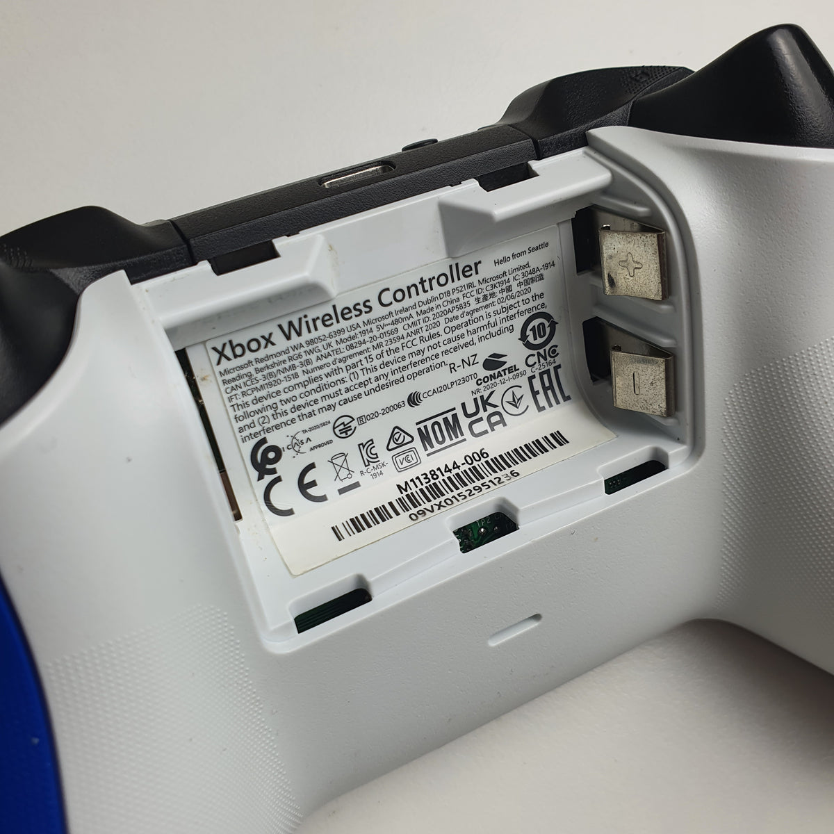 Close-up of the battery compartment of a 'Shock Blue' Xbox wireless controller showing the printed product label, and battery terminals.