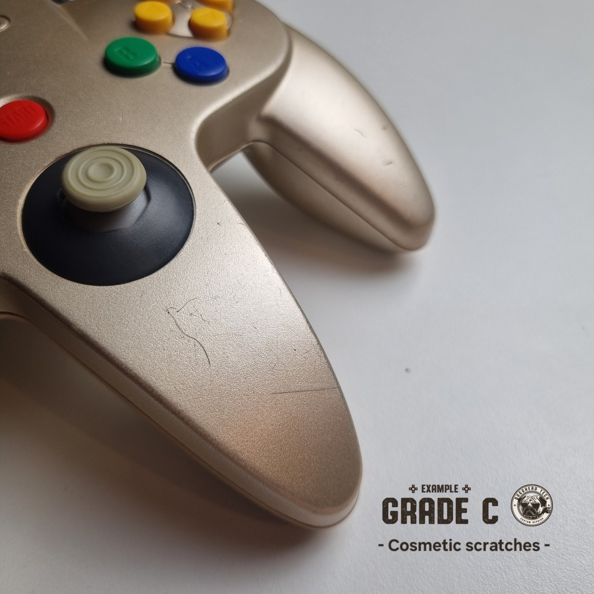 Close-up of a bottom front part of a refurbished modified gold Nintendo 64 controller, showing scratches. The text reads 'Example Grade C: Cosmetic sctratches'.
