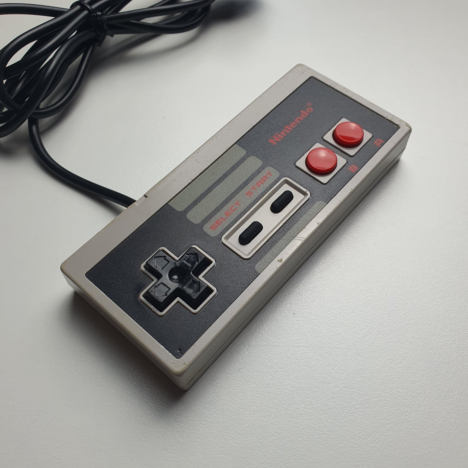 A classic Nintendo NES game controller in grey with red buttons, shown resting on a white surface with its black power cable visible.