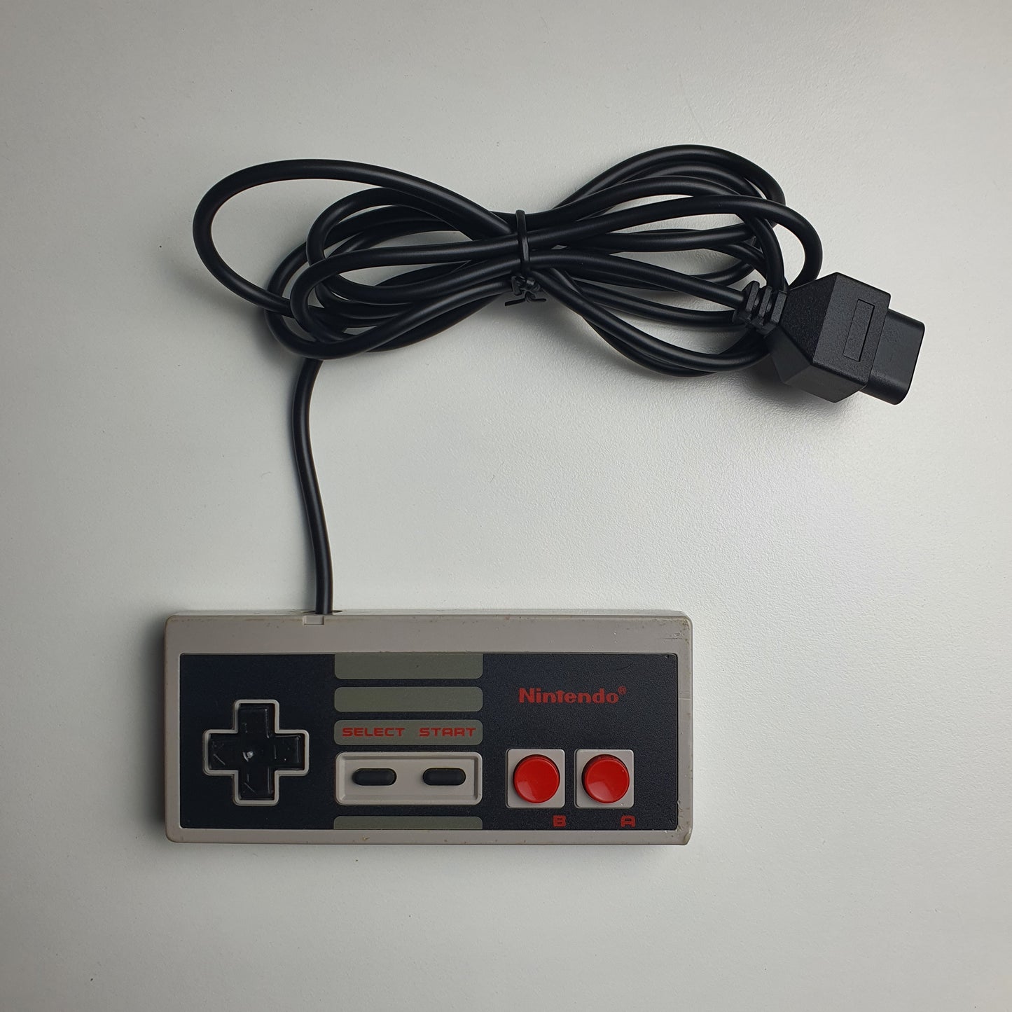 A classic Nintendo Entertainment System NES wired controller with a black coiled cable, photographed on a white background.
