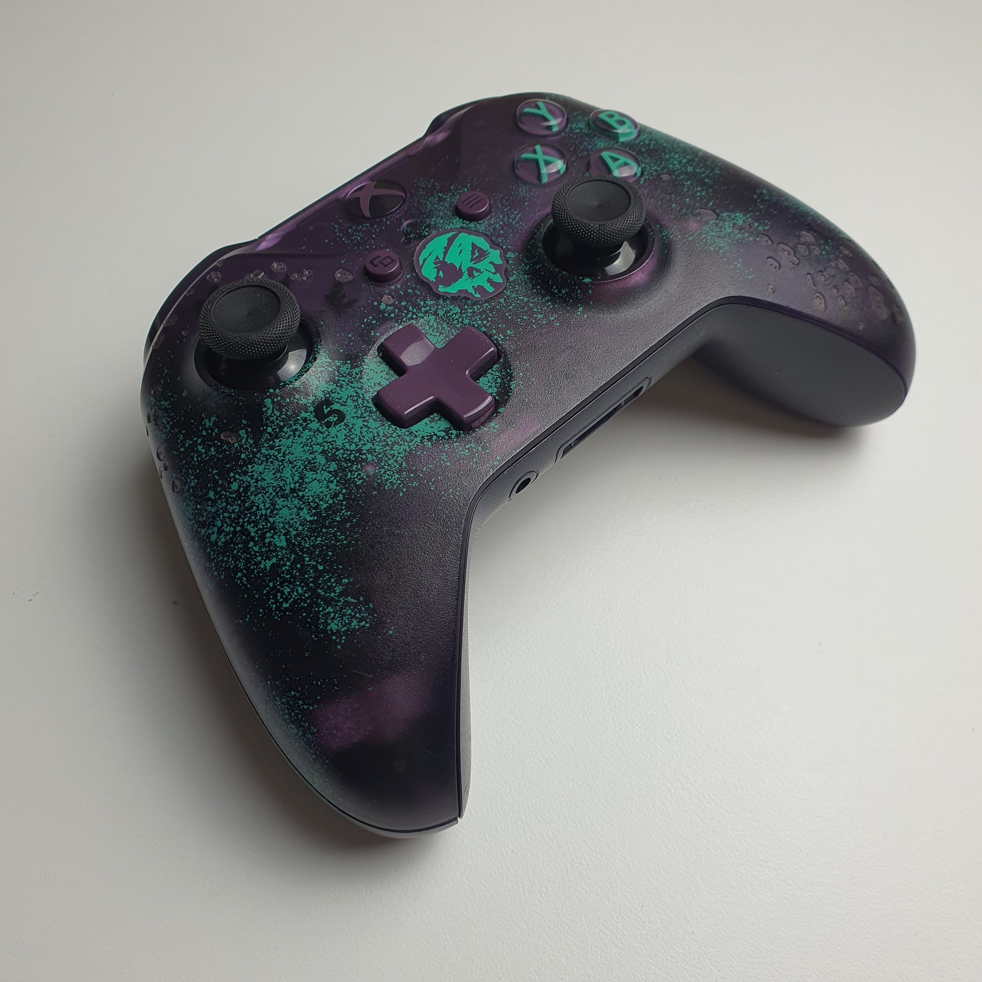 Front of a turquoise & purple Xbox One 'Sea of Thieves' controller on a white background