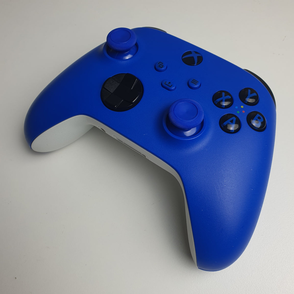 Front of a 'Shock Blue' Xbox game controller showing the front panel, including dual analog sticks, black directional and action buttons, and the Xbox logo, on a light white background.