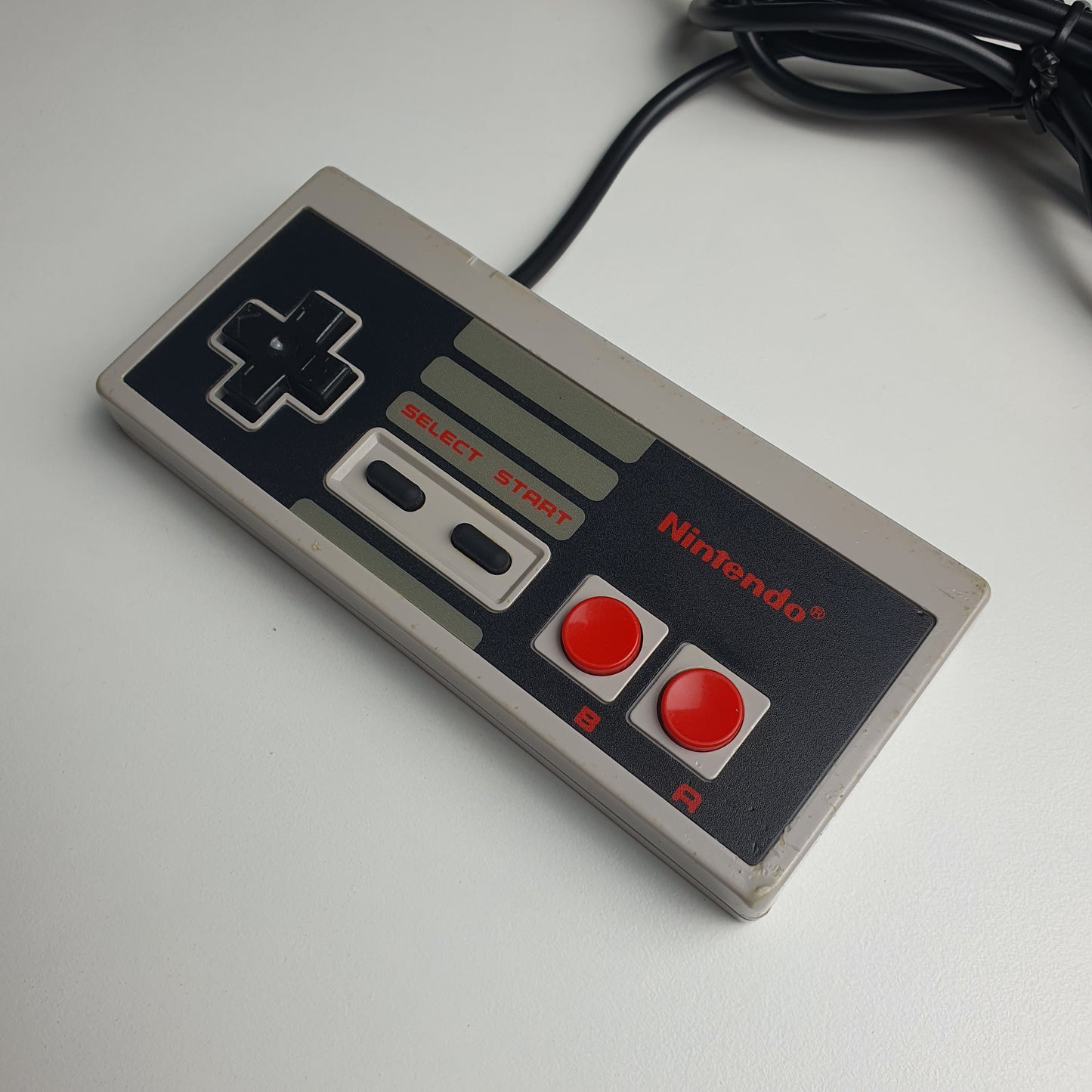 A classic Nintendo NES game controller in grey with red buttons, shown resting on a white surface with its black power cable visible.