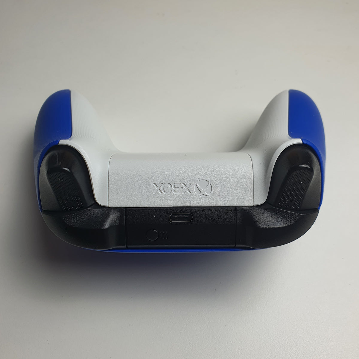 A top-down view of a 'Shock Blue' Xbox gaming controller with a white back, blue side grips, and black textured shoulder buttons, showing the Xbox logo on the battery cover.