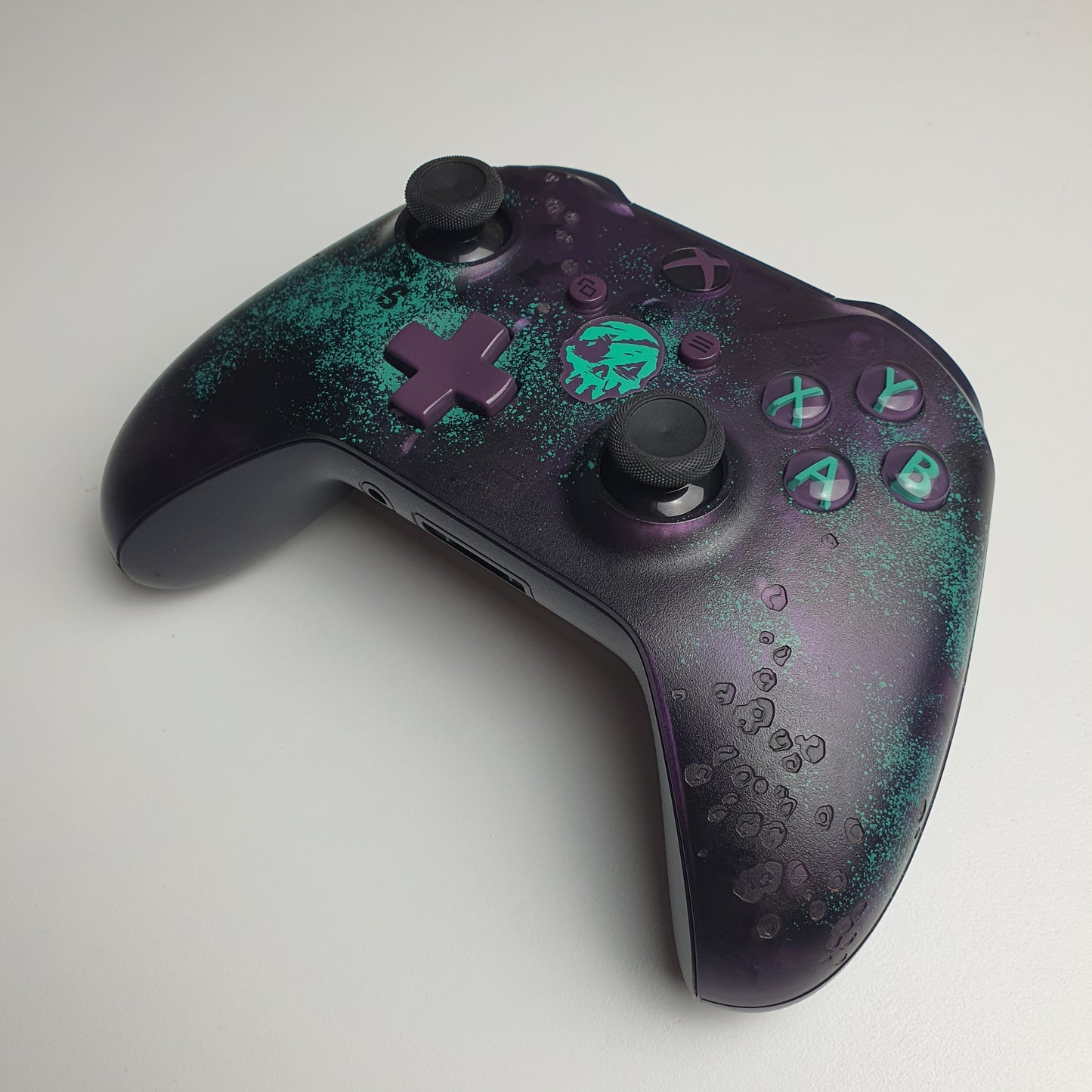 Front of a turquoise & purple Xbox One 'Sea of Thieves' controller on a white background