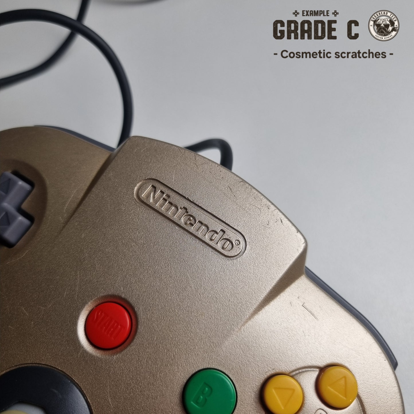 Close-up of a top front part of a refurbished modified gold Nintendo 64 controller, showing scratches. The text reads 'Example Grade C: Cosmetic sctratches'.