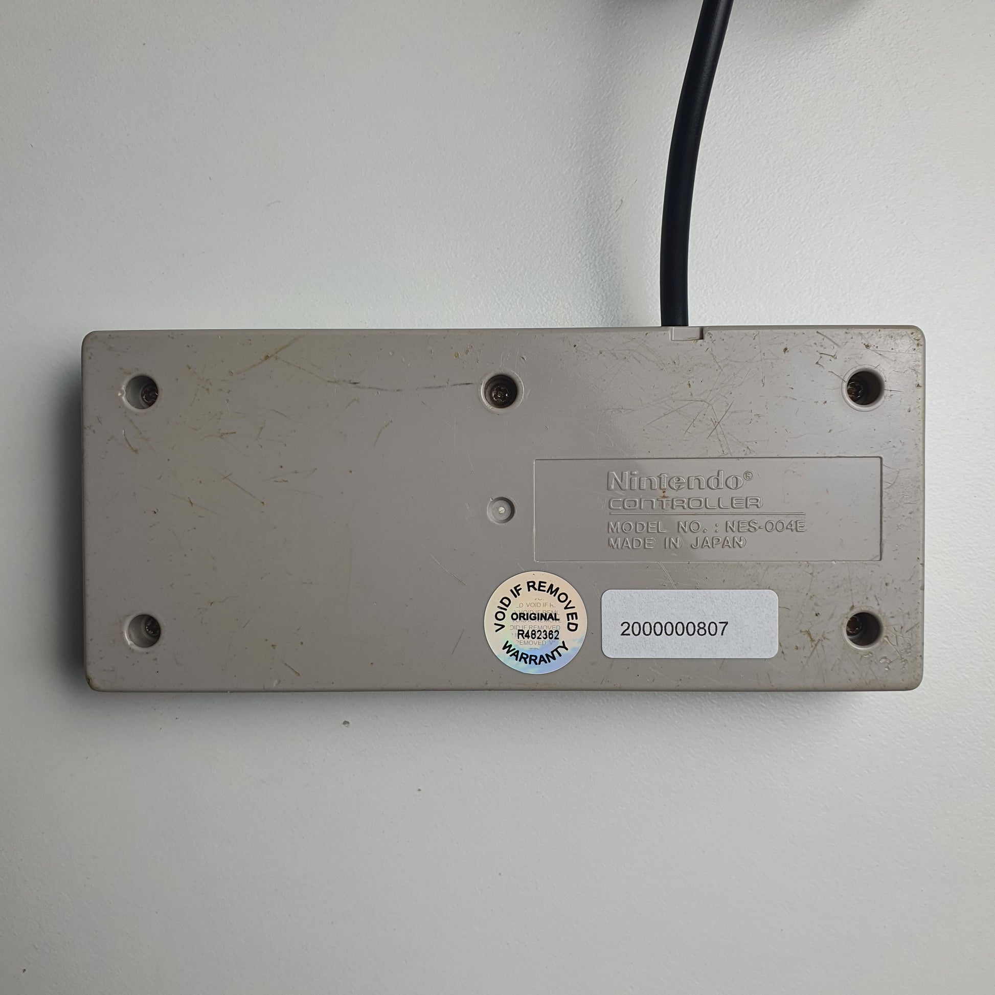The back of a classic Nintendo NES game controller, shown resting on a white surface with its black power cable visible.