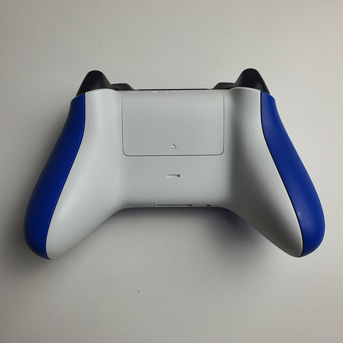 The back of a 'Shock Blue' Xbox game controller showing the white rear panel and battery cover, and blue side panels, on a light white background.